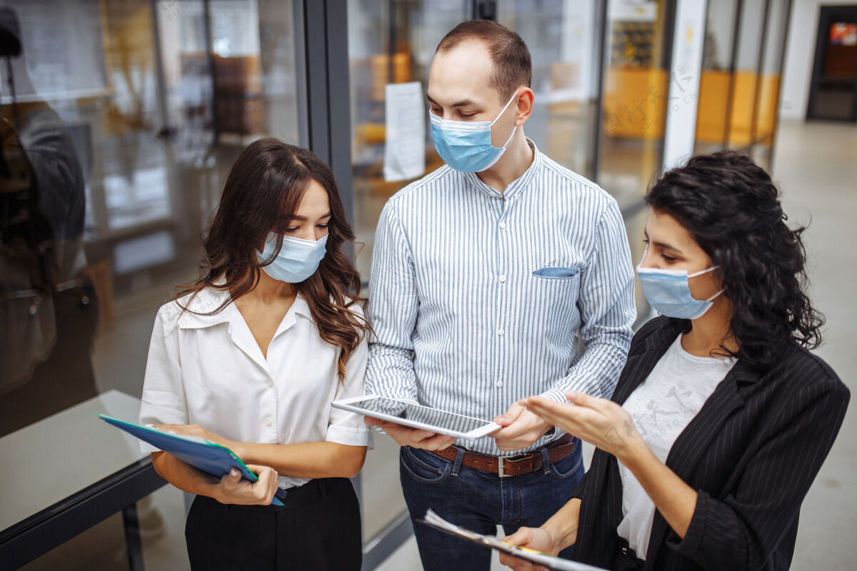 工作三个戴着医用口罩的同事在冠状病毒大流行隔离期间在办公室走廊讨论业务流行病感染工作场所