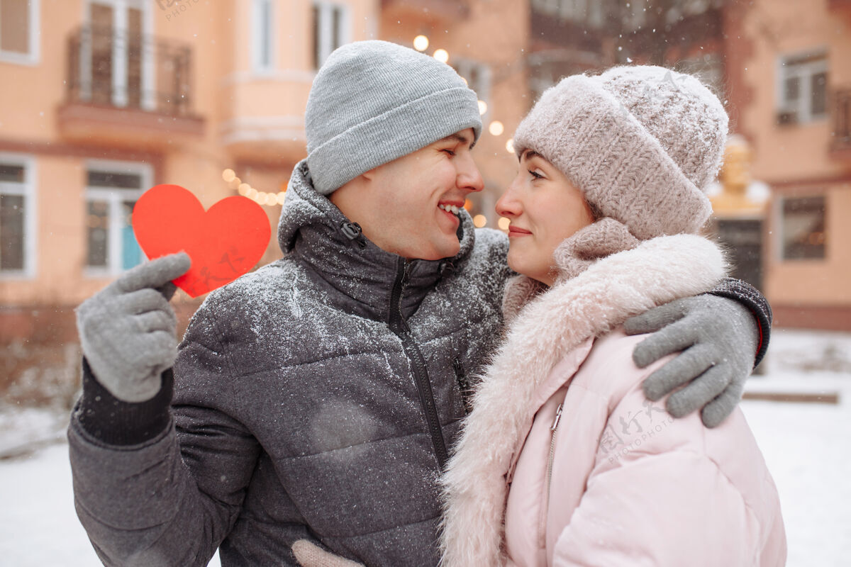 手情人节的概念 相爱的情侣在冬天的雪地里亲吻拥抱帕克·杨男人拿着一个红色的纸心和他的女朋友一起庆祝情人节一对情侣在一起感觉很温暖享受快乐女性