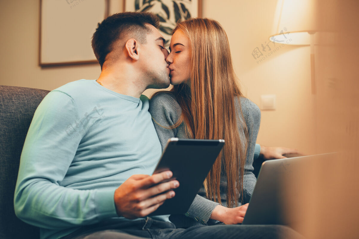 数码一对年轻漂亮的情侣在家里抱着笔记本电脑和桌子 倚在沙发上亲吻观看年轻成人
