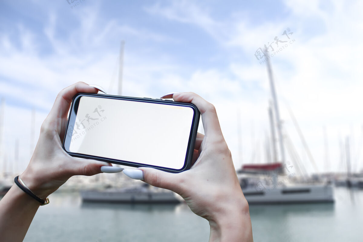 手女孩手里拿着一个智能手机特写镜头 背景是大海和大海游艇.mock-升级技术应用程序日落船