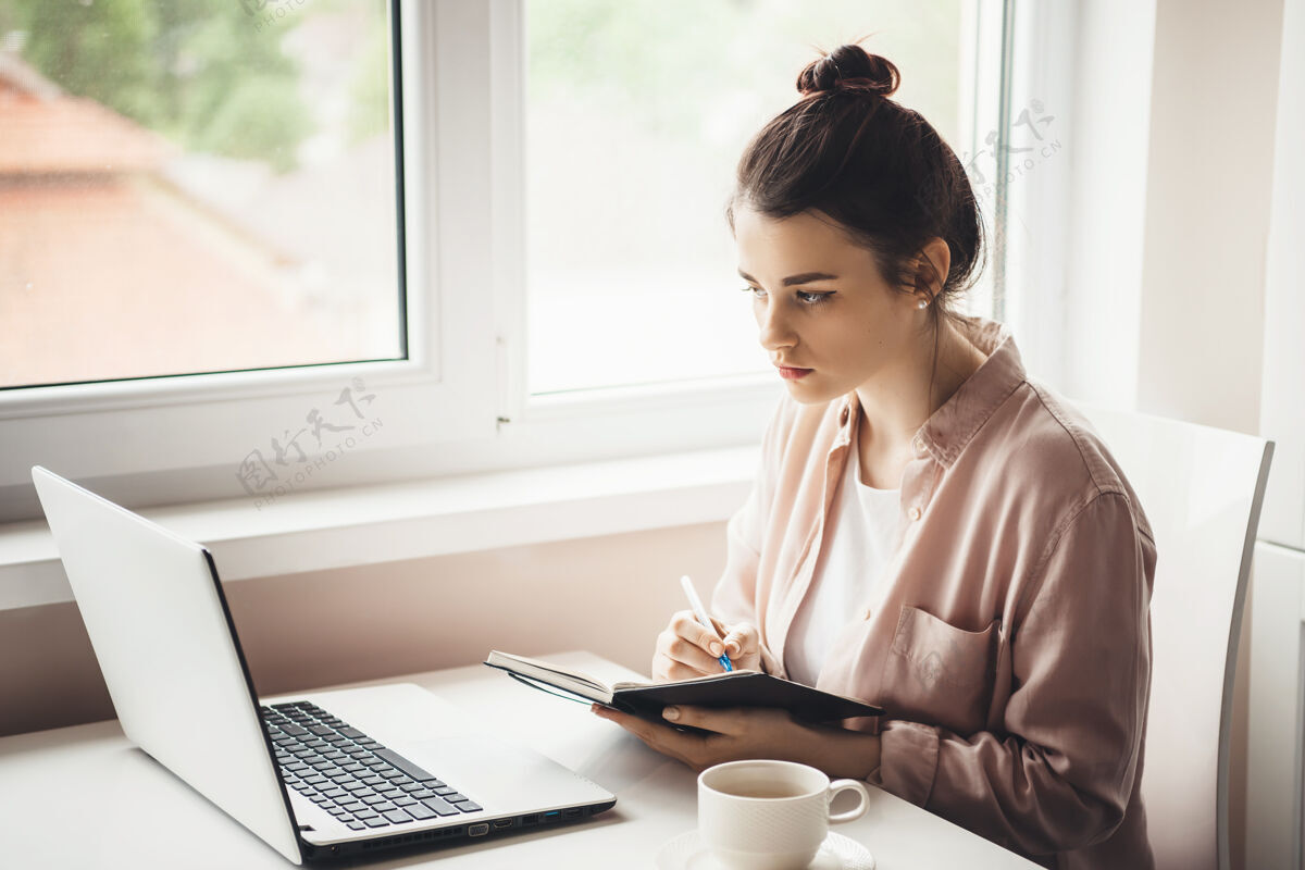 工人一个穿着衬衫的高加索女人一边看着笔记本电脑 一边在窗户边喝茶 一边在书上写食谱在线书房考试