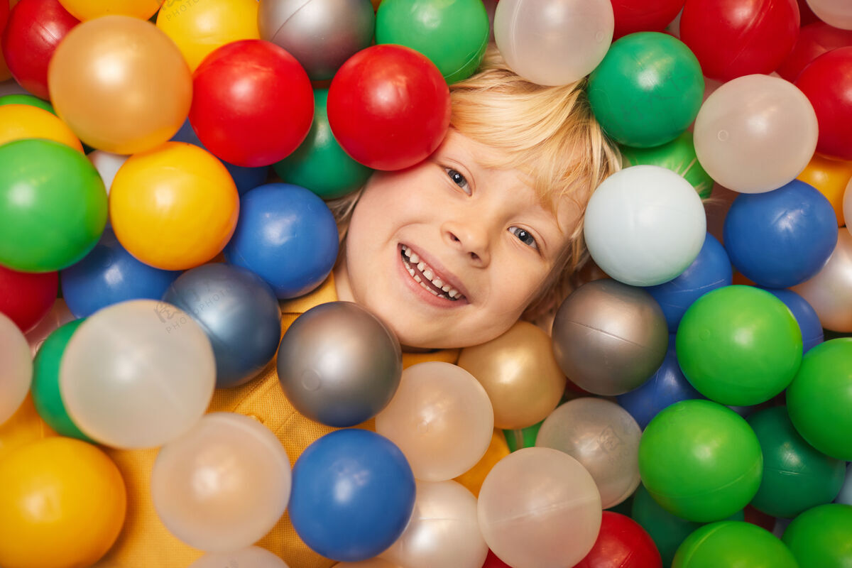 金发金色头发的快乐男孩躺在彩球中玩耍时微笑在前面的特写镜头享受微笑小学