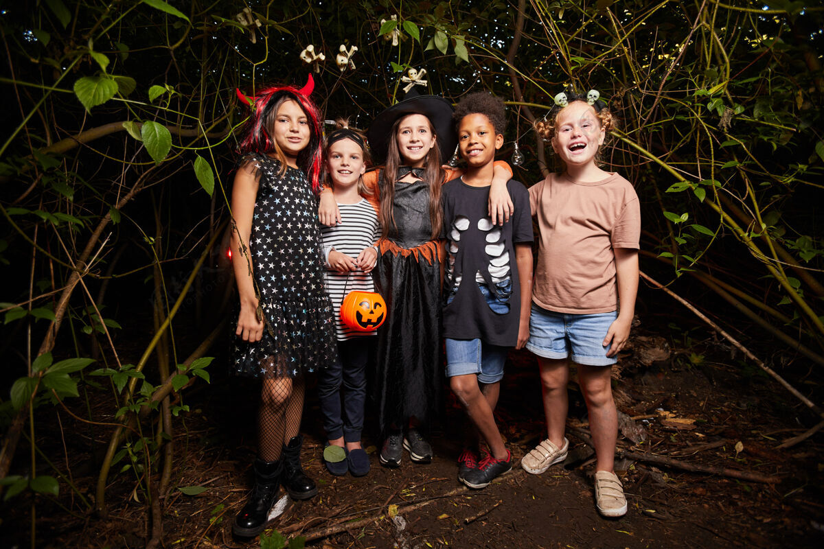 南瓜身着万圣节服装的快乐孩子们站在黑暗的森林里对着镜头微笑的画像夜晚十月聚会社交活动
