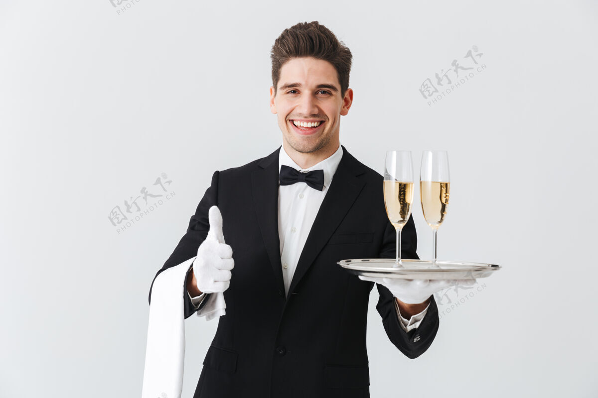 帅气一个身着燕尾服的微笑的年轻侍者的画像在白色的墙上为你提供一杯香槟 竖起大拇指前面饮料杯子