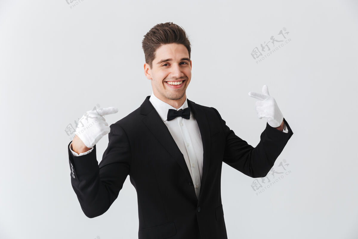 男性一位身穿燕尾服 戴着手套的帅哥侍者孤立地站在白墙上 指着自己的画像鞠躬工业正式服装