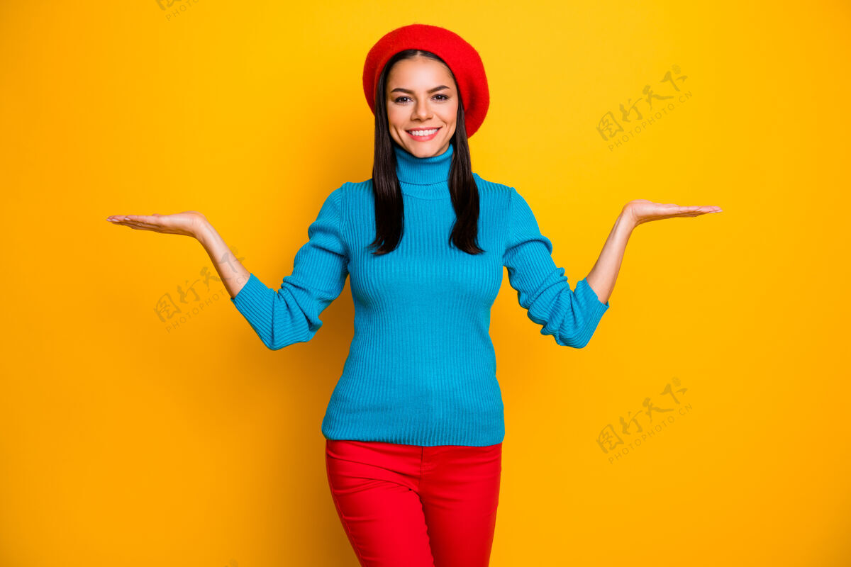 裤子照片中令人惊叹的拉丁女士喜气洋洋的微笑握着张开的手掌新奇的产品戴贝雷帽蓝色高领毛衣红色裤子隔离明亮的黄色墙壁提供选项规模