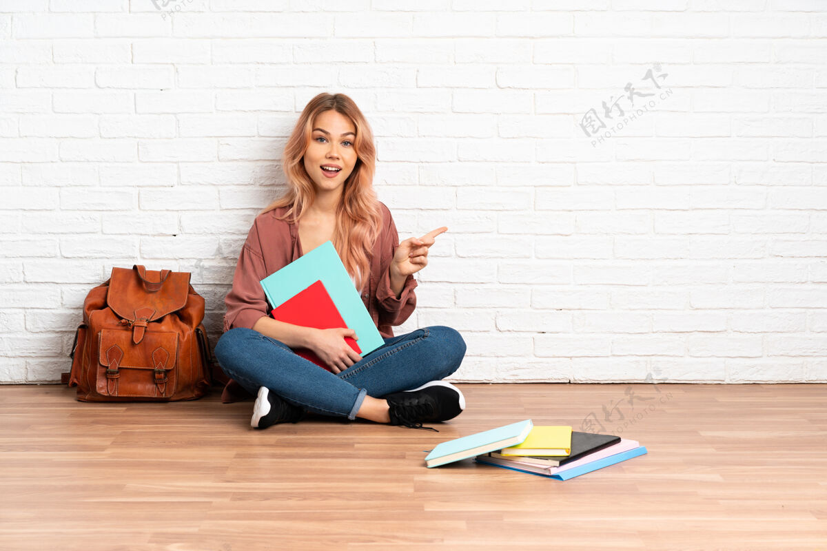 笔记本坐在室内地板上的粉红头发的女学生指着侧面展示产品手指教育肖像