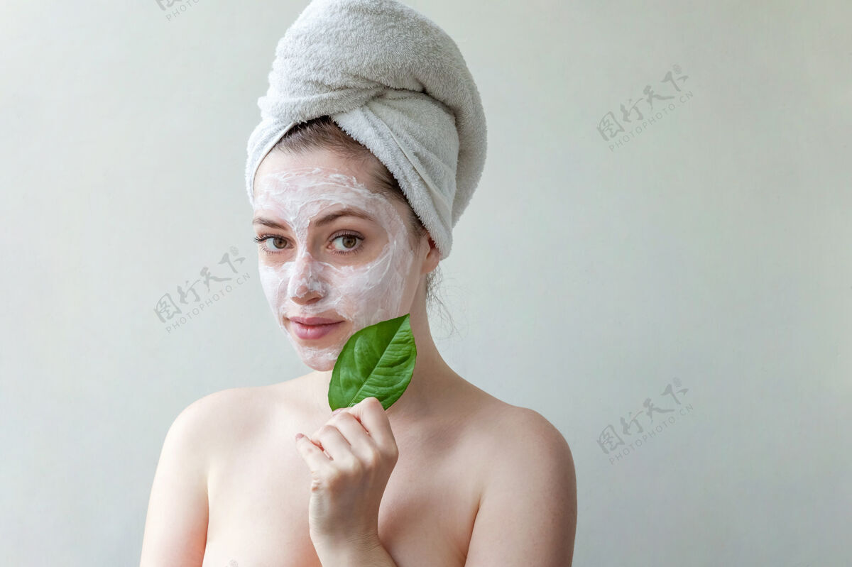 清洁头戴毛巾 脸上涂有白色滋养面膜或乳霜 手拿绿叶 白色背景的美女肖像纯年轻肤色