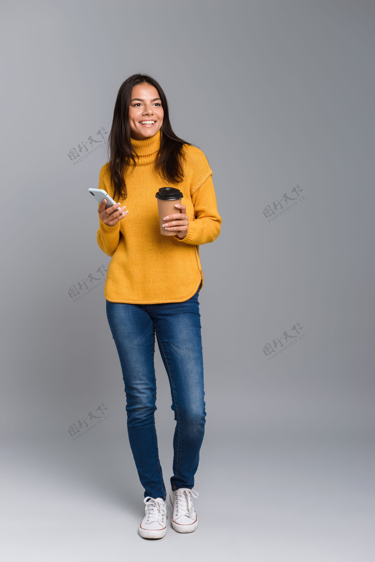 长相一个随意的年轻女子的全长肖像 隔着灰色的墙 拿着外卖咖啡 用手机快乐成人专业