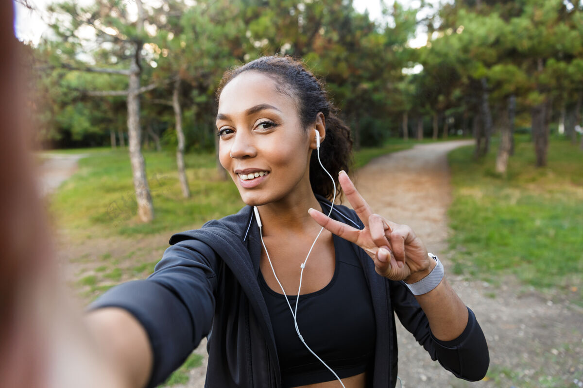 户外20多岁的女性穿着黑色运动服 戴着耳机 在绿色公园散步时用手机自拍女性女人自然