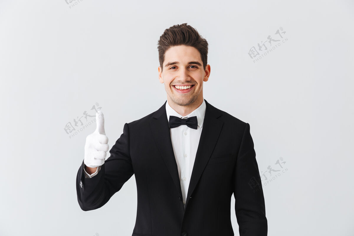 手臂一位身穿燕尾服 戴着手套的帅哥侍者的肖像 孤立地站在白墙上 竖起大拇指餐饮服务员正式服装