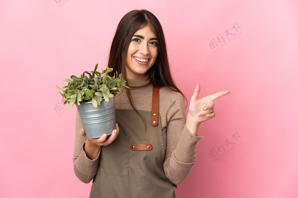 人年轻的园丁女孩拿着粉红色背景上孤立的植物 手指指向侧面笑介绍植物学