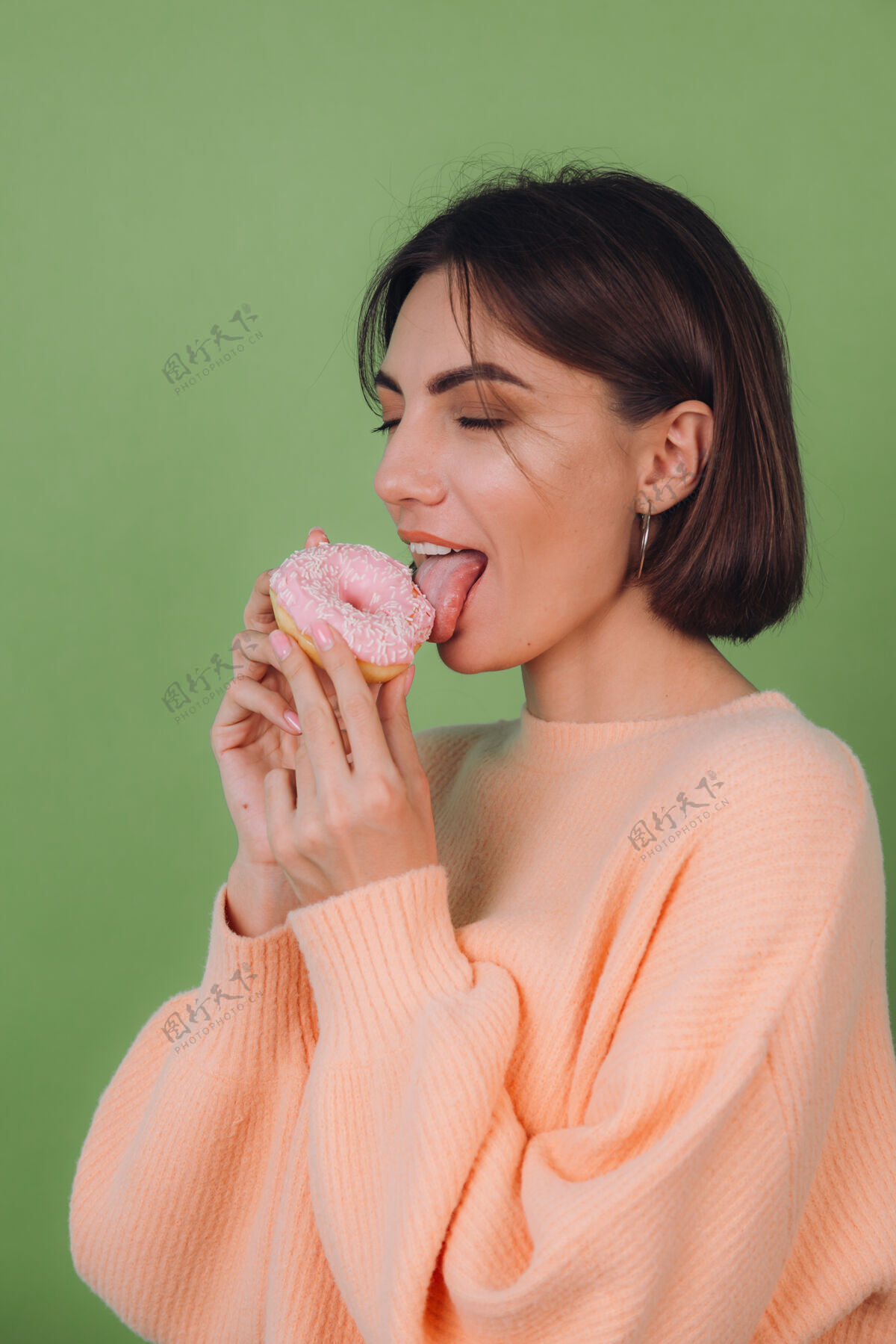 早餐年轻时尚的女人穿着休闲的桃色毛衣孤立地站在绿橄榄色的墙上舔着粉色的甜甜圈复制空间年轻肖像休闲