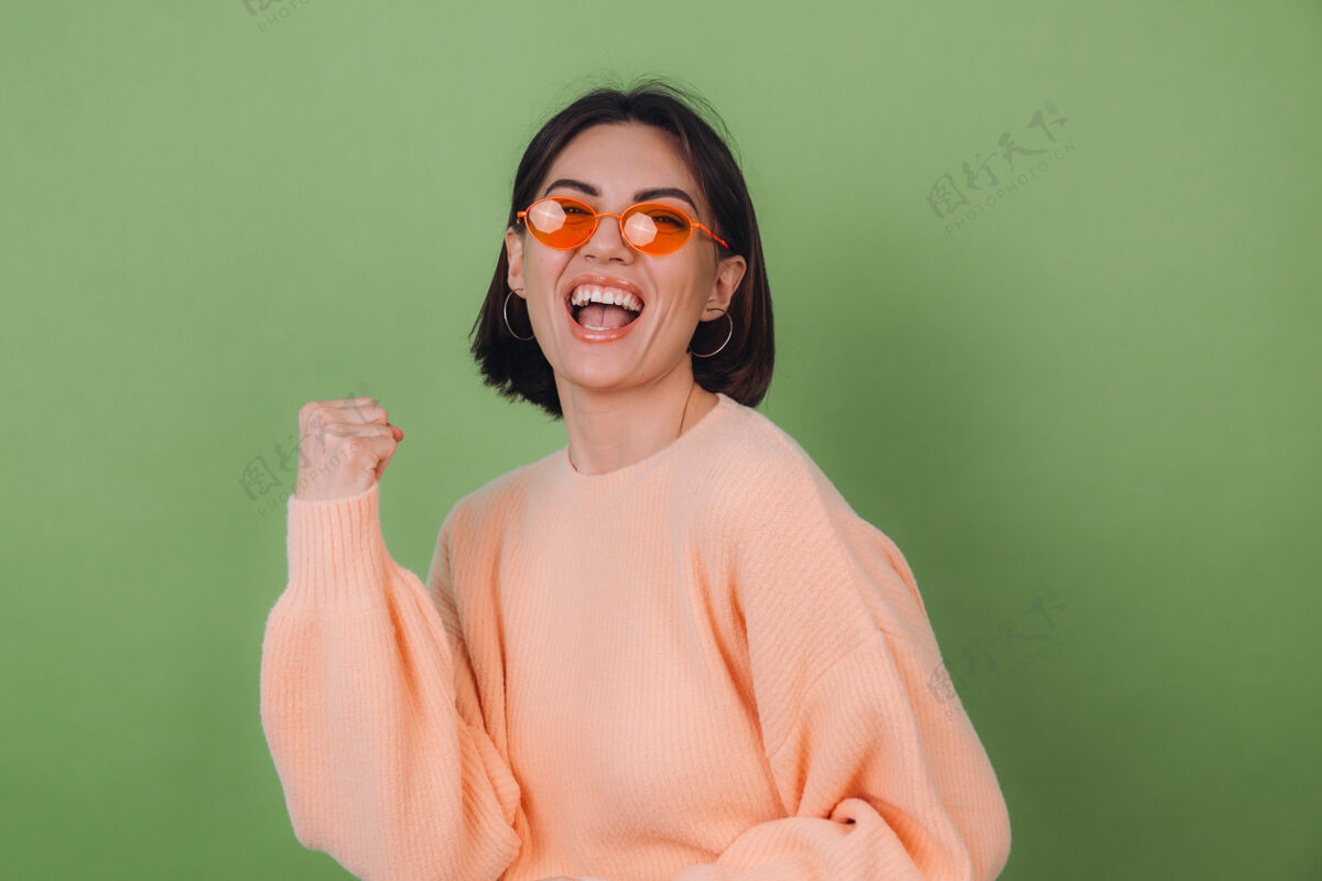 年轻年轻时尚的女士穿着休闲的桃色毛衣 戴着橙色眼镜 隔着绿橄榄色的墙壁积极地做着赢家的手势握紧拳头复制空间人物奇迹黑发