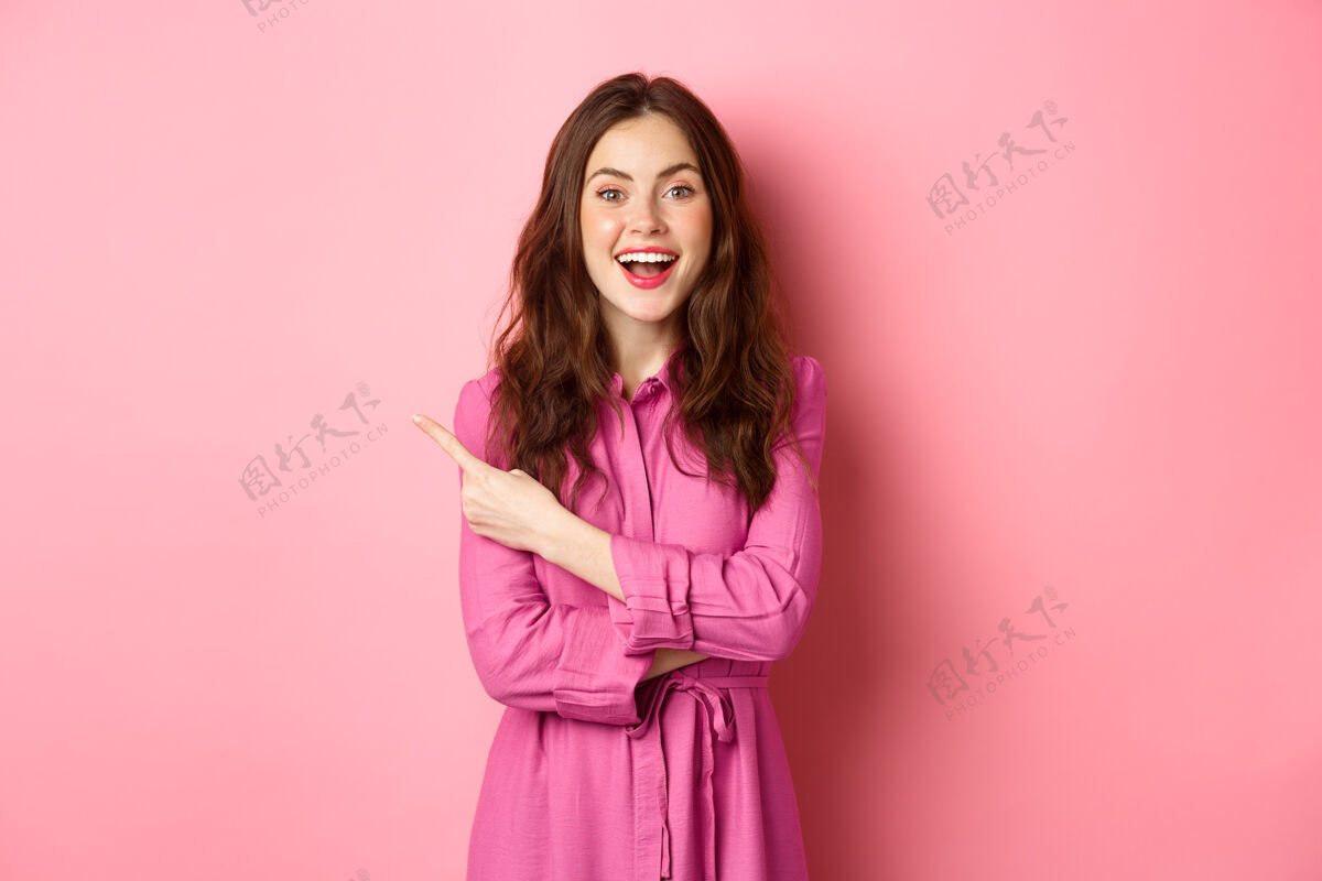 女人时尚的年轻现代女性看起来很惊讶 手指放在左边的文字空间 展示宣传文字 幸福地站在粉红色的墙上一旁情绪候选人