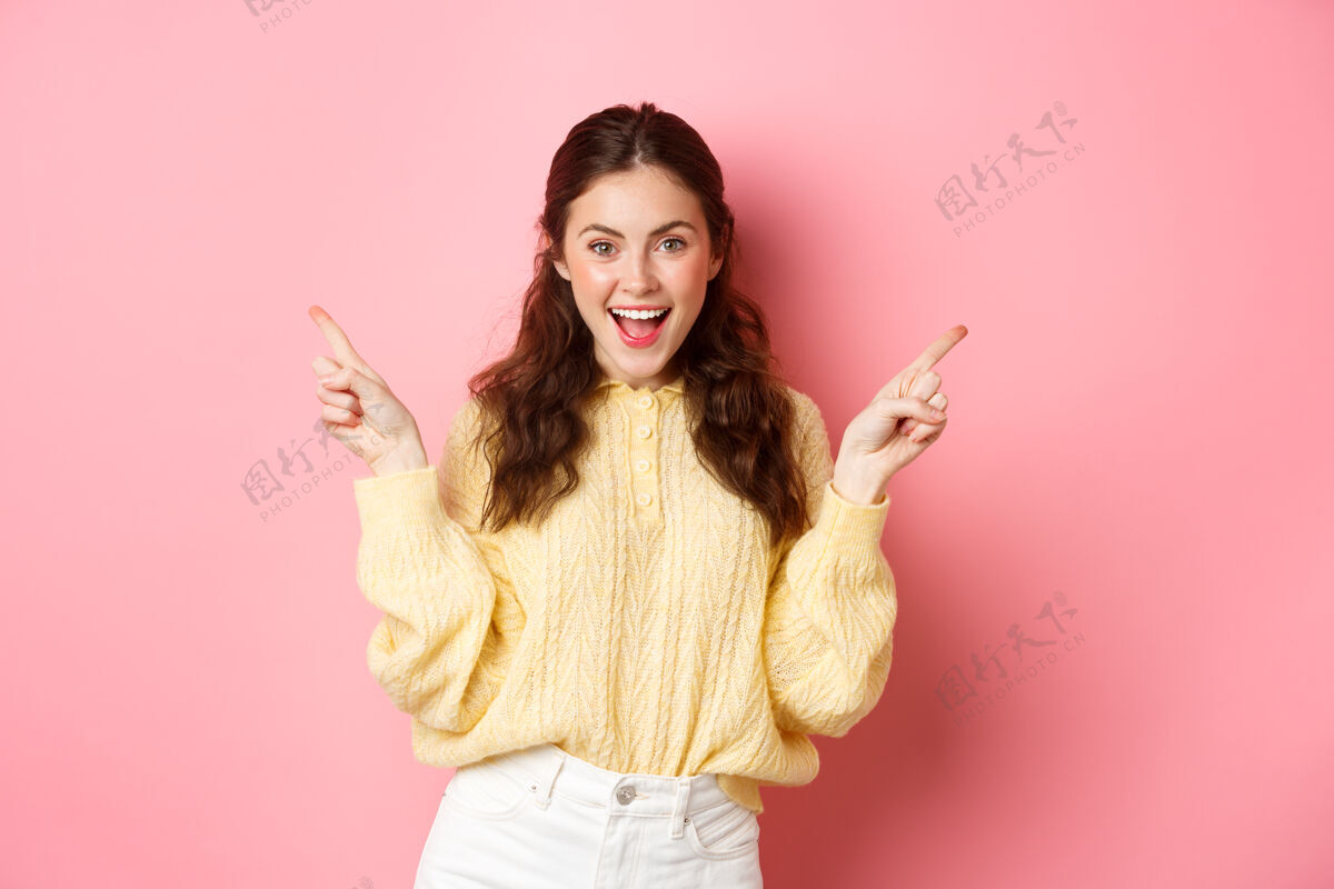 休闲一个开朗漂亮的女模特把手指放在一边 展示了两个选择 左边和右边的标志 站在粉红色的墙上欢呼魅力一旁