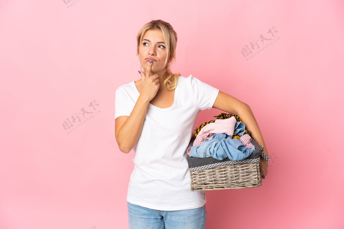 俄罗斯一个年轻的俄罗斯女人拿着一个衣服篮子 孤立地站在粉红色的背景上 一边抬头一边怀疑衣服沉思洗衣