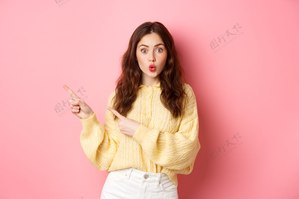 时尚惊讶的黑发女子展示促销短信 喘气逗乐了 手指指着左边的促销 站在粉红色的墙上兴奋魅力女人
