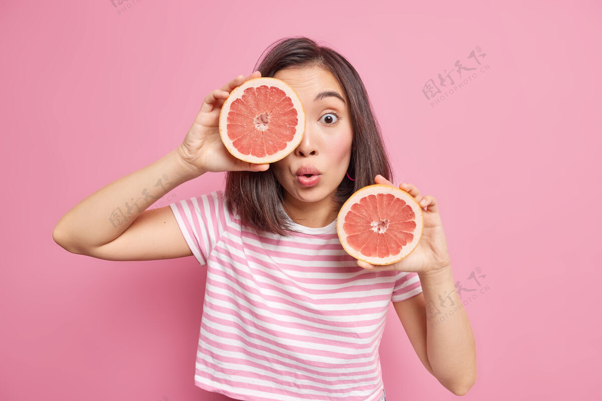 素食者惊讶的亚洲年轻女子在室内摆出柑橘类水果的姿势 手里拿着两半新鲜的柚子 保持健康的营养 穿着t恤衫 表情震惊 隔着粉色的墙壁柑橘皮肤模特
