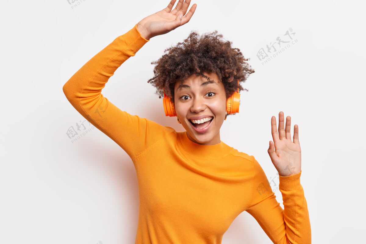 年轻快乐快乐的非洲裔美国妇女享受令人敬畏的音质戴立体声无线耳机听喜爱的音乐穿着橙色套头衫在白色墙壁上的节奏舞蹈隔离女士个人手掌