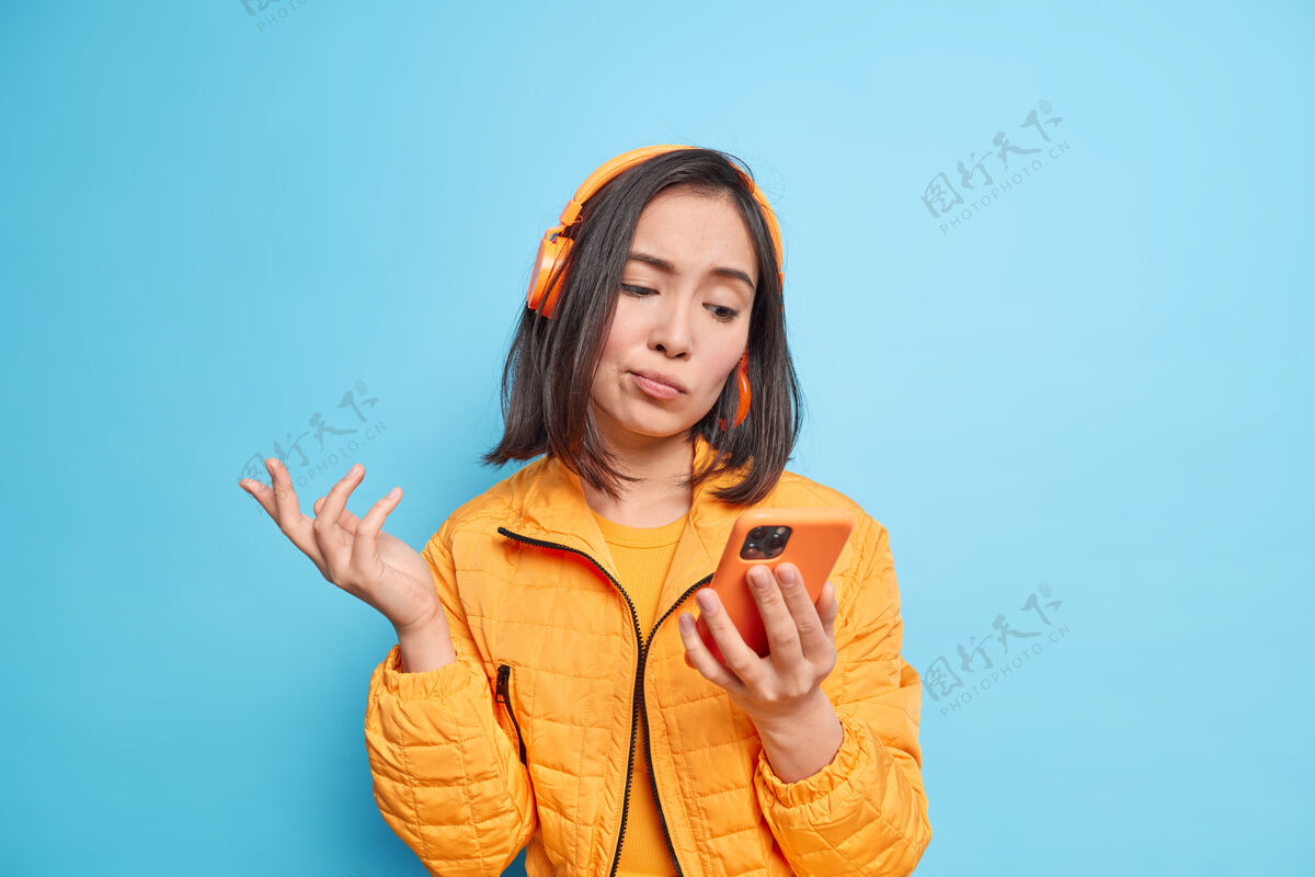 心烦迷惑不解犹豫不决的亚洲女人看着智能手机屏幕不能选择歌曲听戴着无线耳机耳朵穿着时尚夹克隔着蓝色的墙焦点学生女