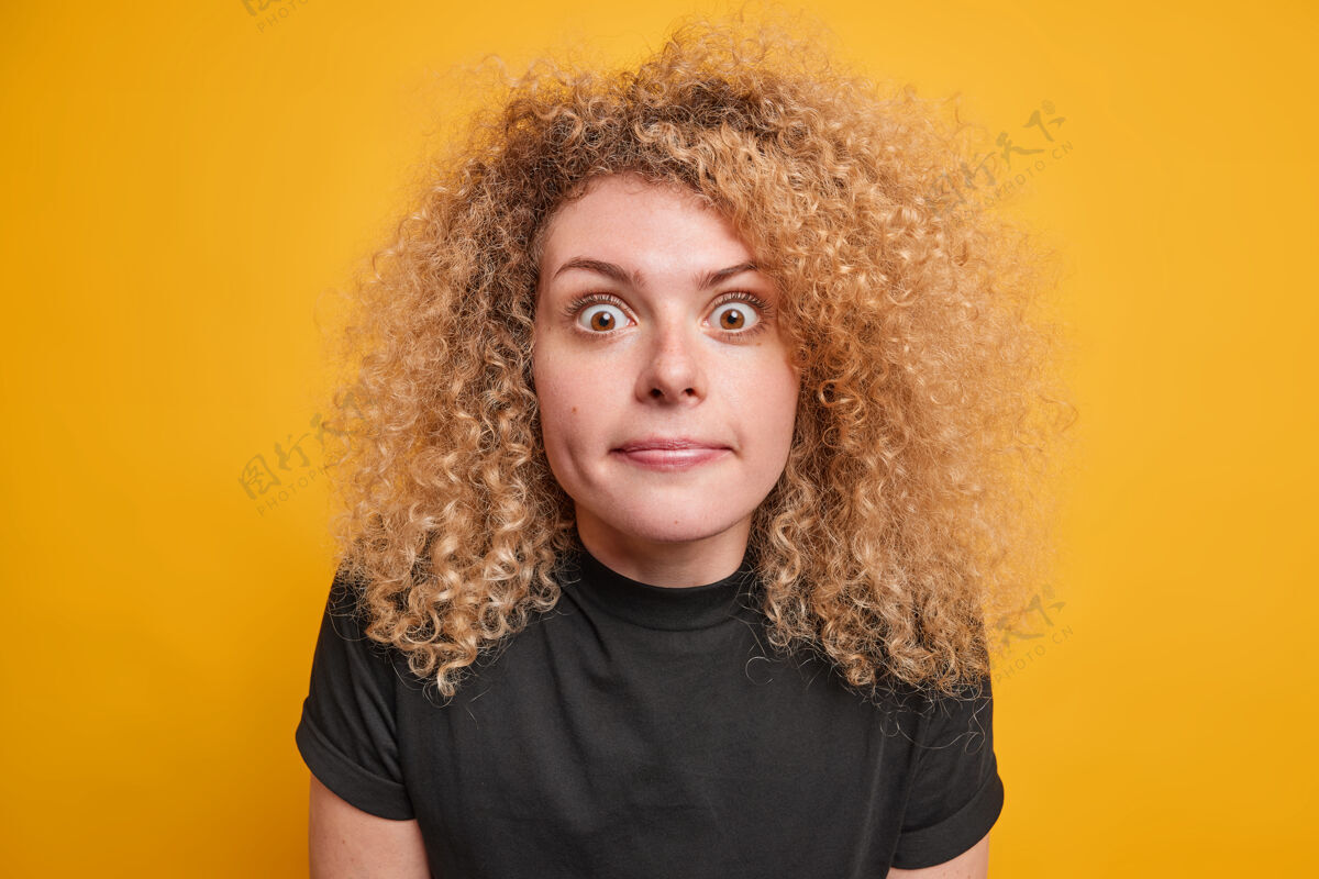 自然一个自然卷发的年轻欧洲女人穿着休闲的黑色t恤 隔着黄色的墙壁 惊讶地对一些奇怪的表情做出反应女性惊呆随意