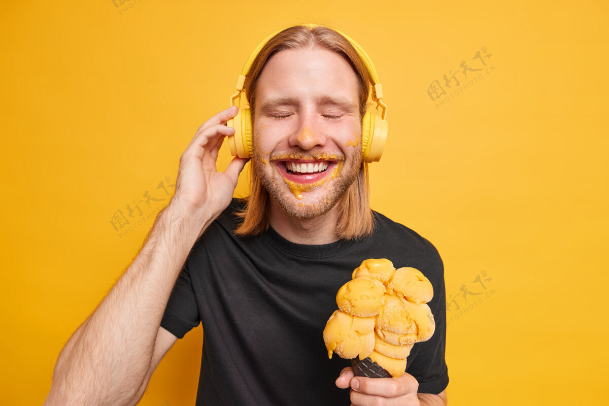 表情正高兴的红发小伙闭上眼睛 开心地笑着 喜欢的歌 手戴着耳机 手里拿着美味的大冰激凌 有空余的时间 在夏天的一天休息 隔离在黄色的墙上脏请男性