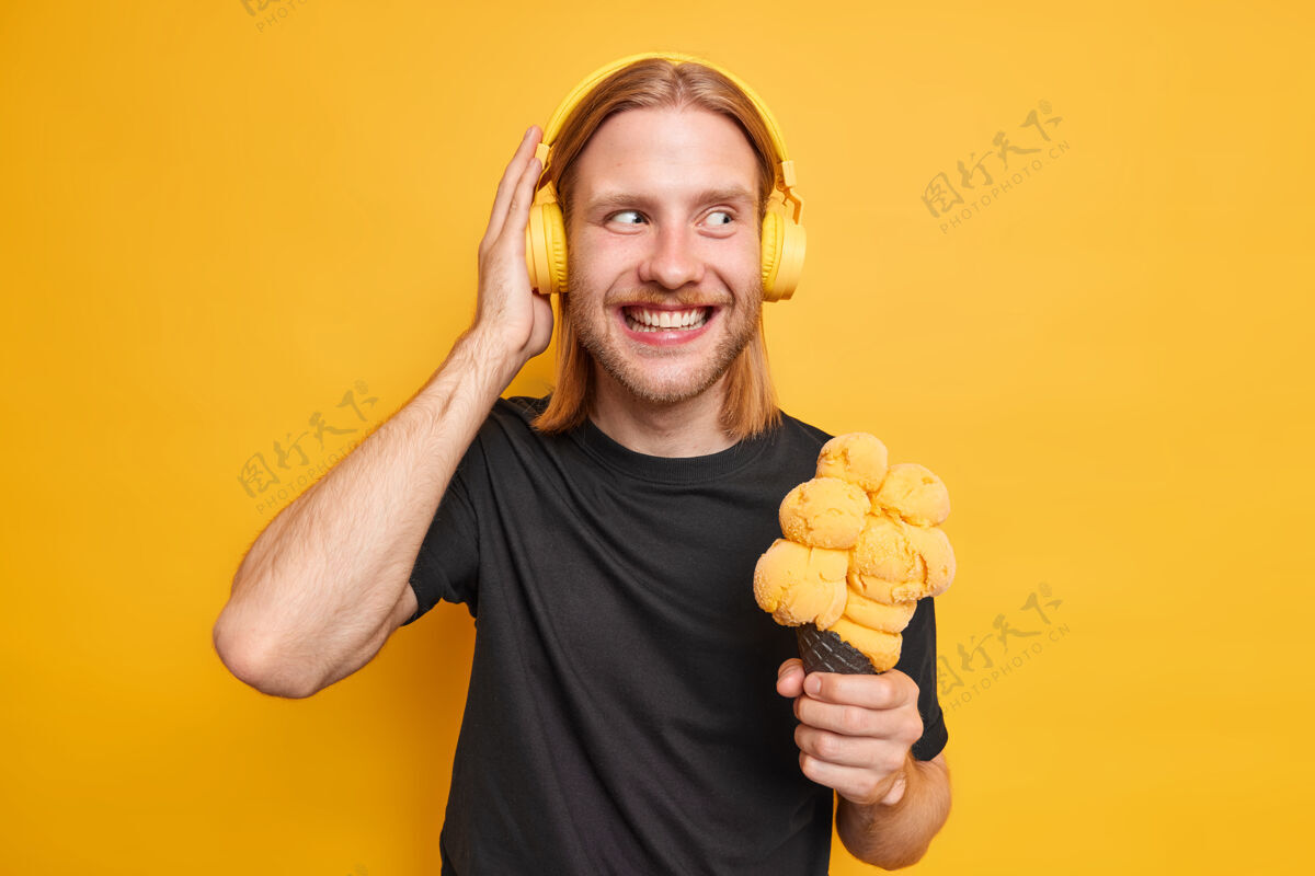 单人积极的红胡子男人手放在耳机上拿着美味的冰激凌通过耳机享受喜爱的音乐穿着随意隔离在黄色的墙壁时髦的家伙与冰淇淋冰淇淋手势冰