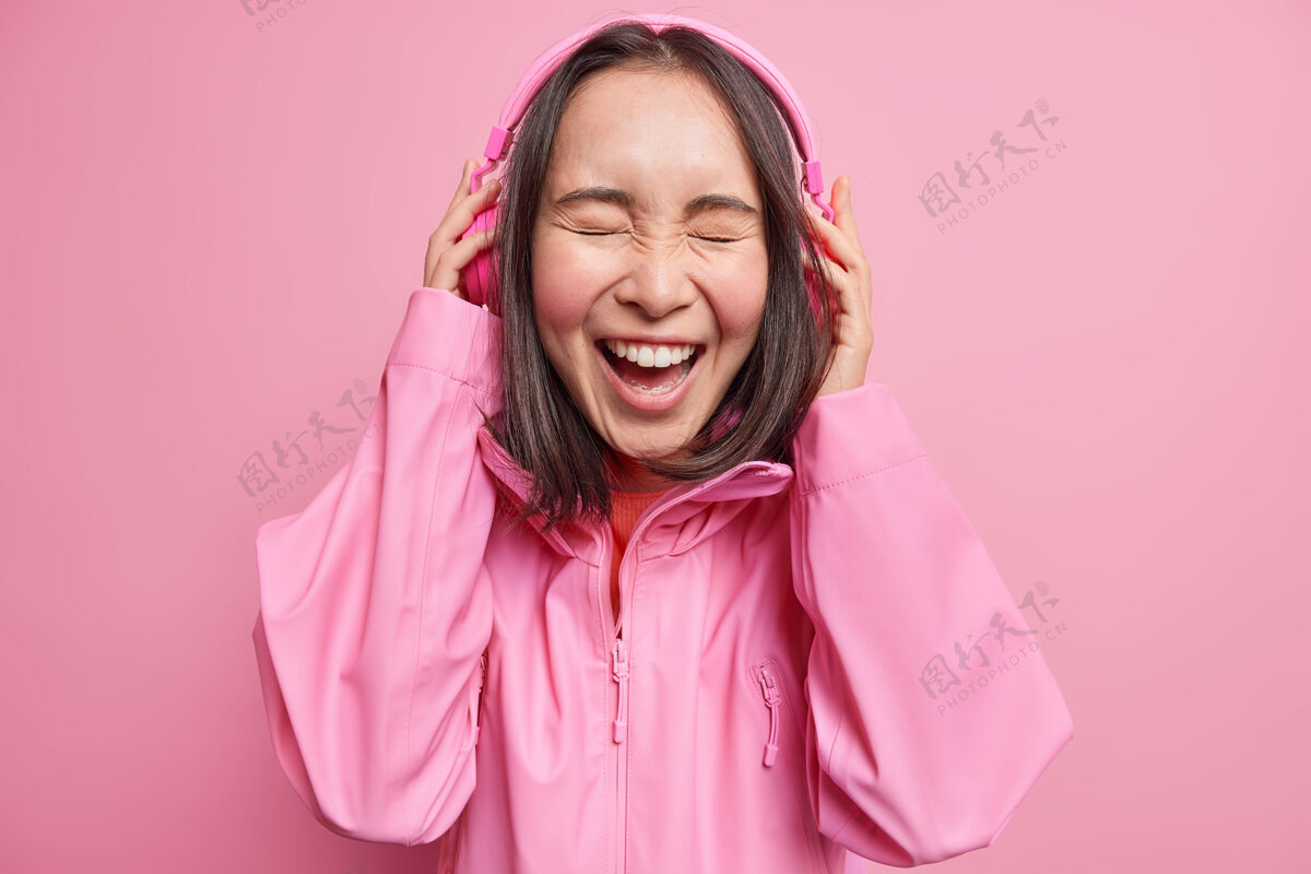 女性美丽的黑发亚洲女人通过立体声耳机在线听有趣的故事笑着开心地闭上眼睛从喜悦穿夹克隔离在粉红色的墙壁人们的情感生活方式的概念成人可爱魅力