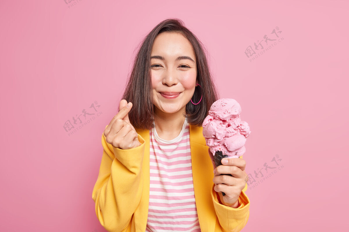 女高兴的亚洲女人做出迷你心形韩式手势拿着美味的冰淇淋吃着甜甜可口的冷冻甜点穿着条纹t恤黄色夹克对着粉色的墙壁摆姿势夏天到了小吃夏天冷