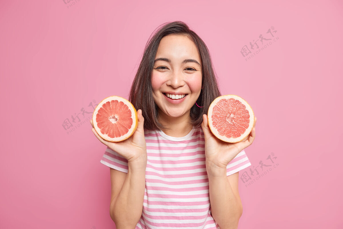 乐观可爱开朗的亚洲女人 深色头发 拿着两片葡萄柚 用柑橘类水果做天然化妆品 穿着随意 有快乐的表情 隔着粉色的墙壁素食人青少年