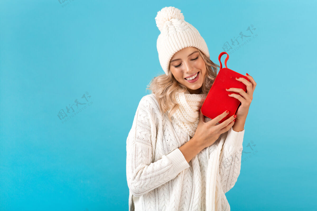 舒适时尚的金发微笑的美丽年轻女子手持无线扬声器听音乐快乐地穿着白色毛衣和针织帽子冬季时尚摆造型穿着女性女士