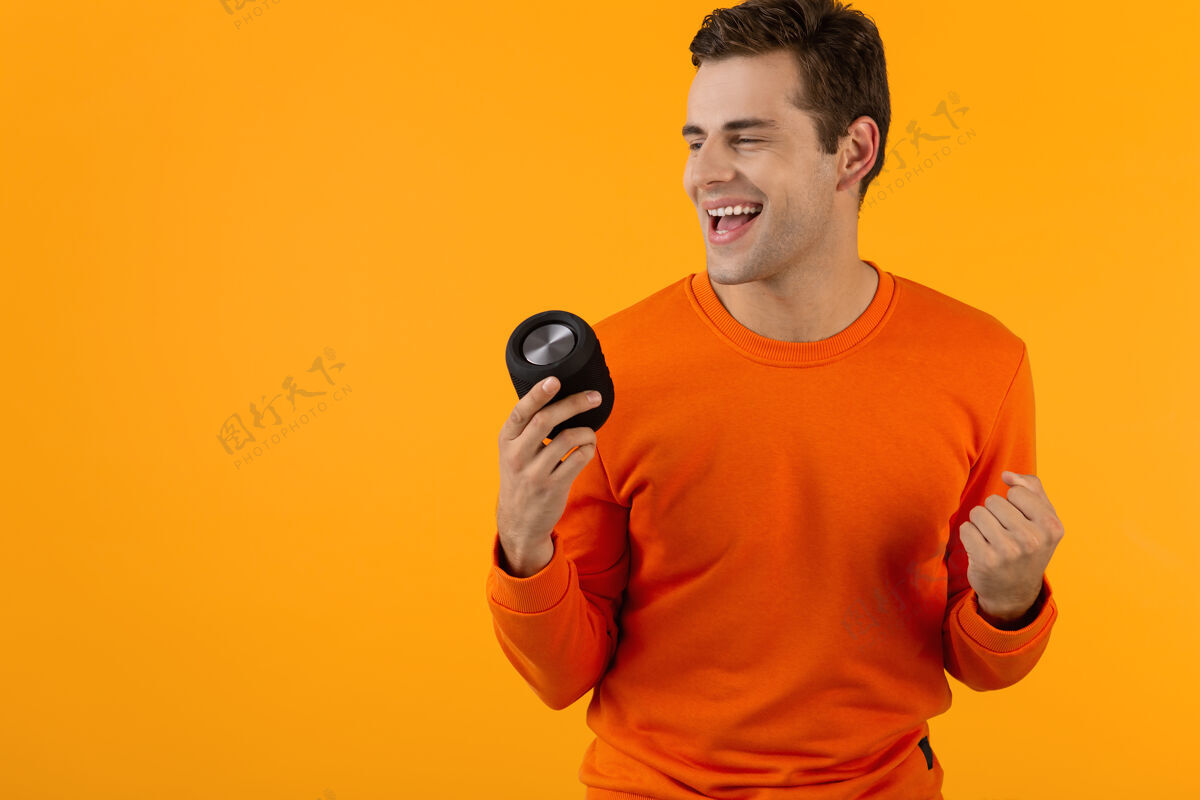 无线时尚微笑的年轻人穿着橙色毛衣手持无线扬声器快乐地听音乐玩得开心欢乐男性时尚