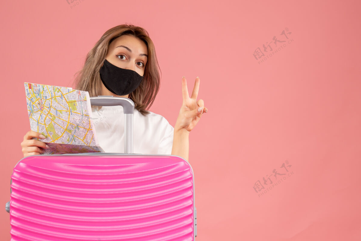 地图正面图：戴着黑色面具的年轻女子拿着地图 在粉色手提箱后面做着胜利的手势面具旅游肖像