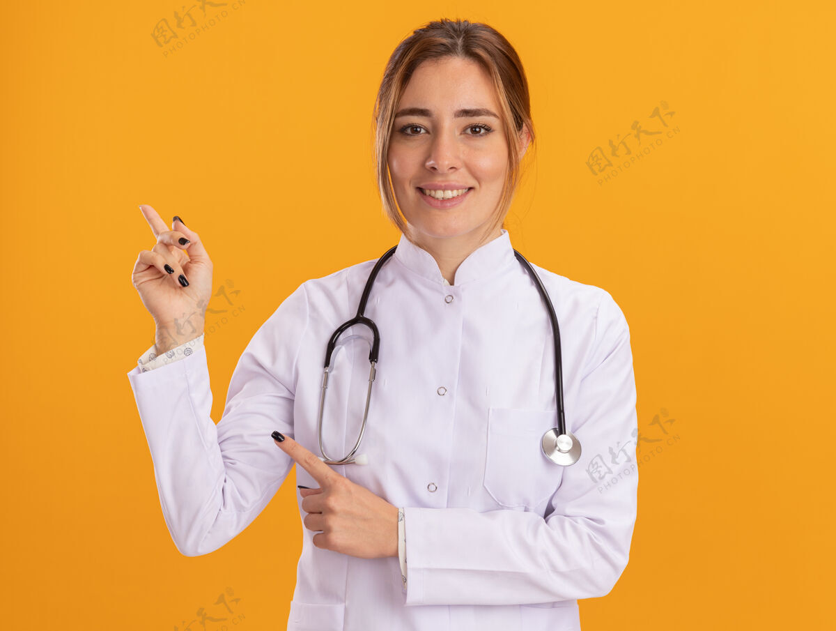 人面带微笑的年轻女医生穿着医用长袍 听诊器指向隔离在黄色墙壁上的一侧 并留有复印空间衣服表情点