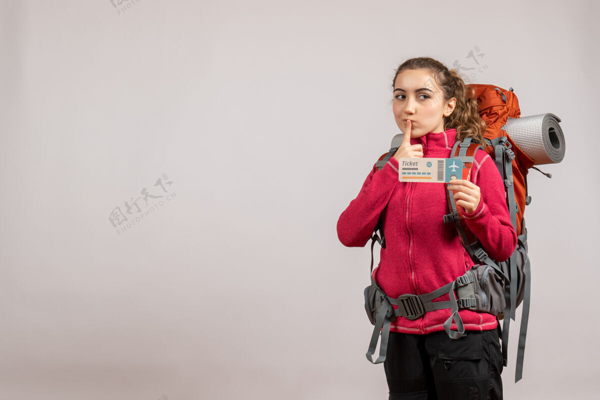 青年正面图：年轻的旅行者背着大背包 举着旅行票 手指放在嘴上冒险票旅游