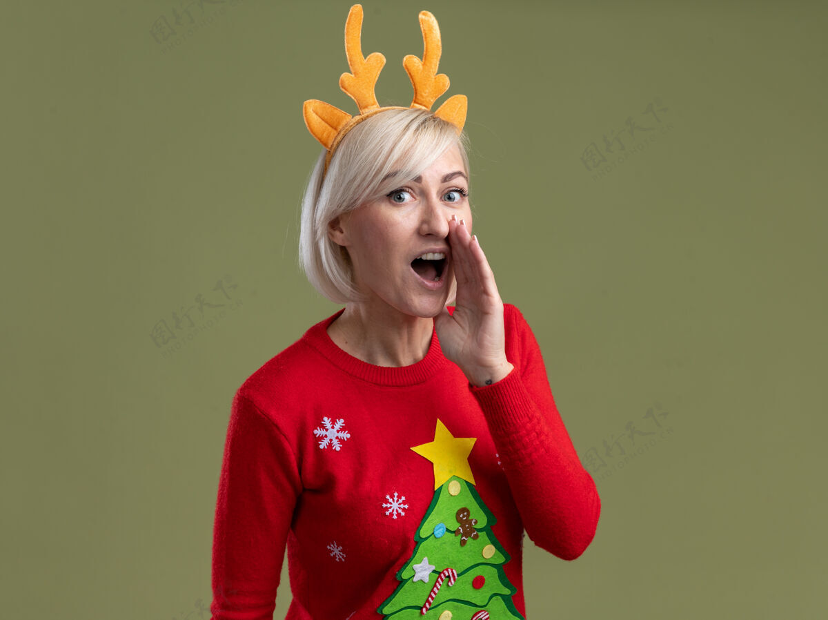 近令人印象深刻的中年金发女人戴着圣诞驯鹿鹿角头带和圣诞毛衣看着保持手靠近嘴窃窃私语隔离在橄榄绿的墙壁与复制空间手女人穿