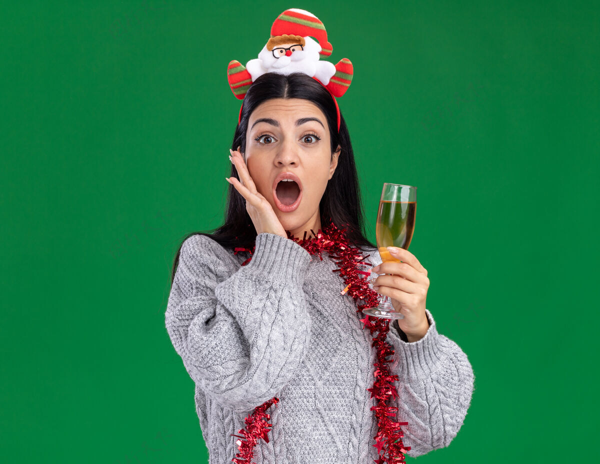穿关心的年轻白人女孩戴着圣诞老人的头带 脖子上戴着金箔花环 手里拿着一杯香槟 手放在脸上 隔离在绿色的墙上 留着复制空间玻璃圣诞老人花环