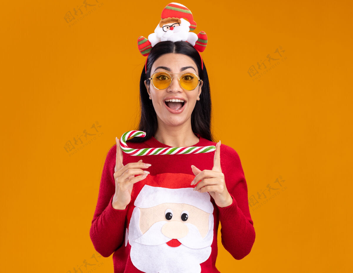 眼镜令人印象深刻的年轻白人女孩戴着圣诞老人的头带和毛衣与眼镜举行传统的圣诞糖果手杖水平隔离在橙色墙上的复制空间圣诞老人水平圣诞节