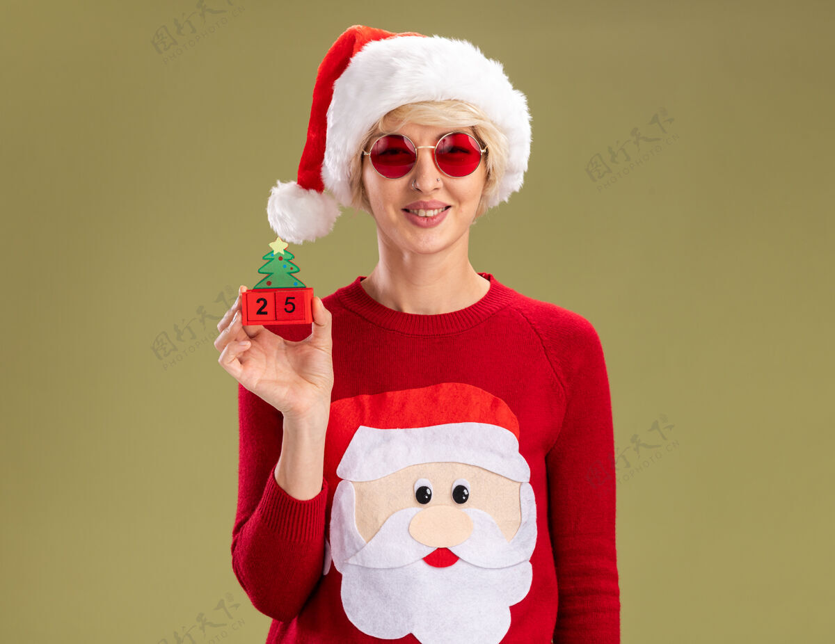 眼镜微笑的金发女郎戴着圣诞帽和圣诞老人的圣诞毛衣戴着眼镜拿着圣诞树玩具和日期看起来孤立在橄榄绿的墙上复制空间橄榄女人金发