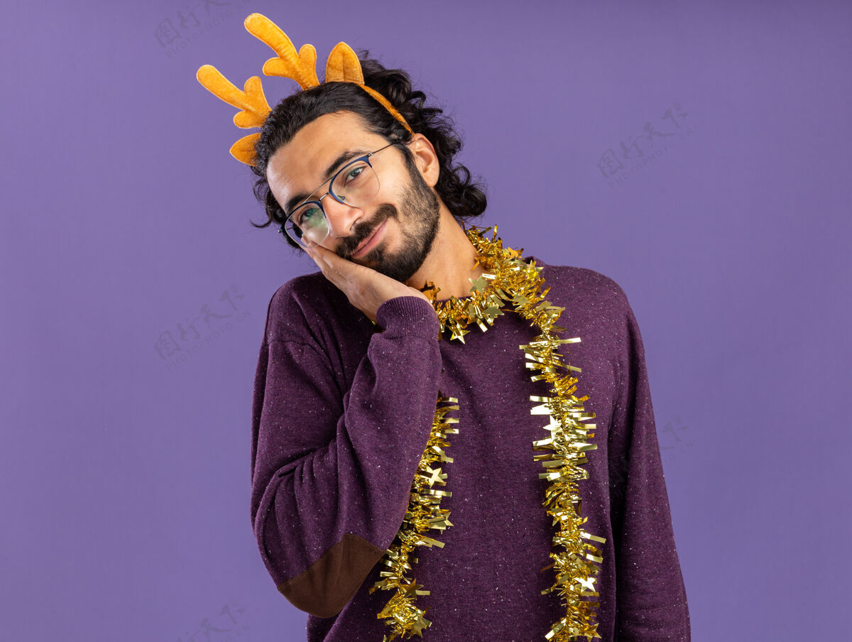 帅哥高兴的歪着头的年轻帅哥戴着圣诞发箍脖子上戴着花环手放在脸颊上隔离在蓝色的墙上花环箍穿