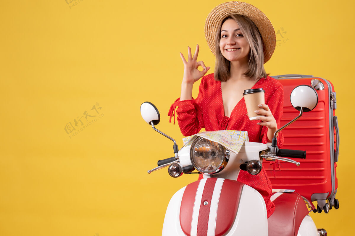 旅游正面图：身穿红色连衣裙的年轻女士拿着咖啡杯 在轻便摩托车旁做着“ok”的手势行李度假持有
