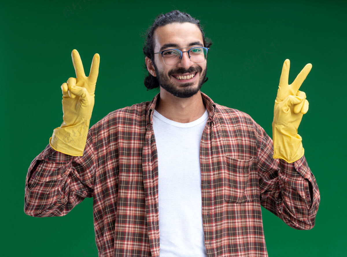 年轻微笑的年轻帅气的清洁工穿着t恤 戴着手套 在绿色的墙上显示出和平的姿态和平手势帅气