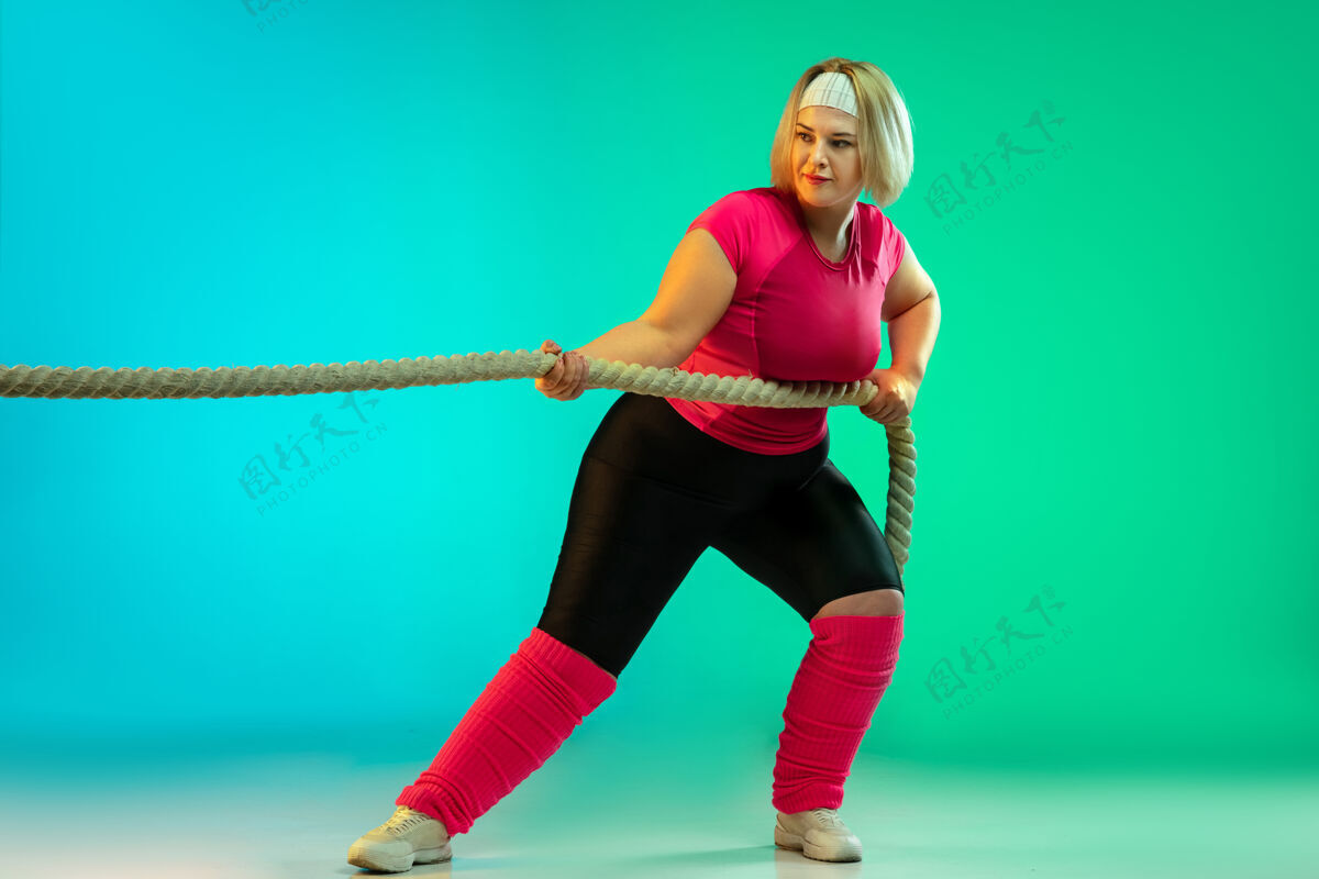 多样性年轻的白人大号女模特在霓虹灯下的渐变绿色背景下训练用绳子做锻炼运动的概念 健康的生活方式 积极的身体 平等女子加上举重