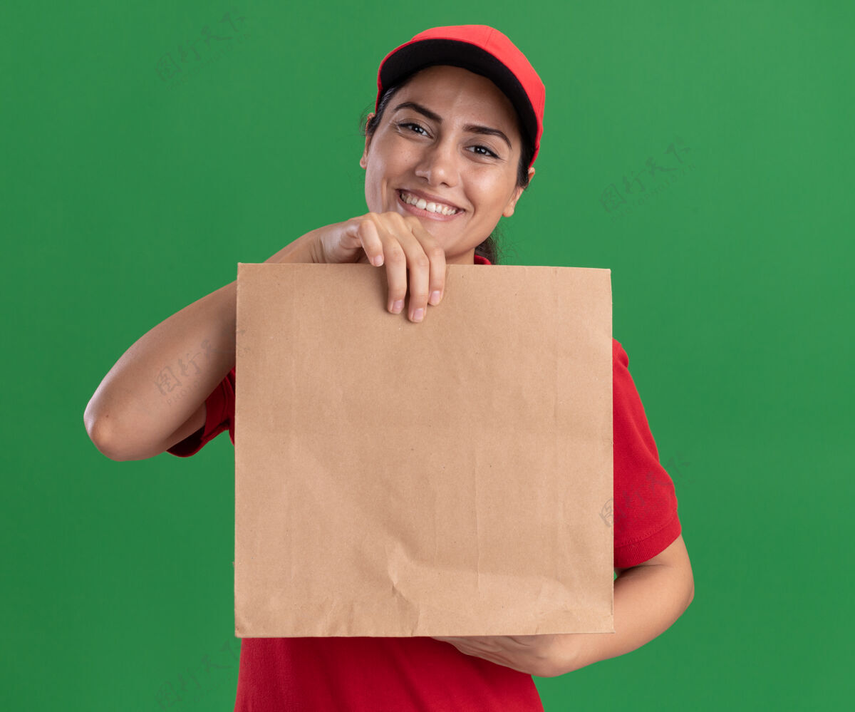 姿势微笑的年轻送货女孩穿着制服 戴着帽子 拿着纸食品包装隔离在绿色的墙上感觉穿着脸