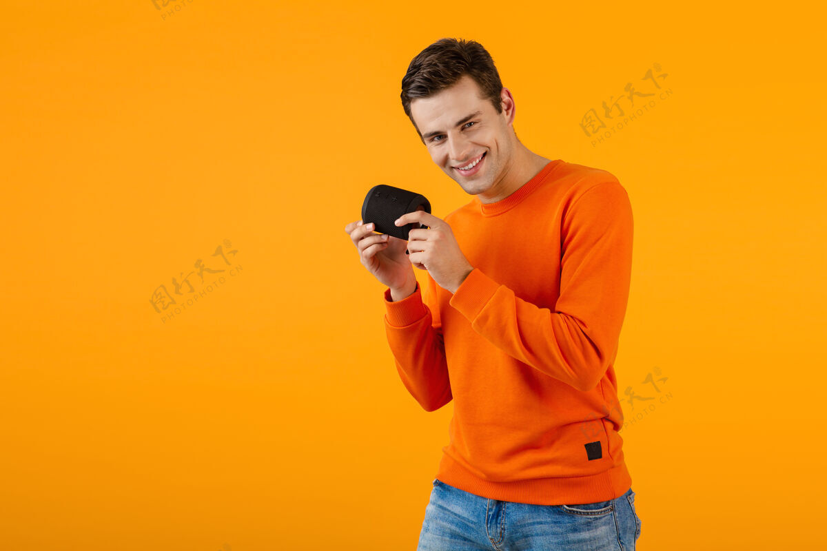 城市时尚微笑的年轻人穿着橙色毛衣手持无线扬声器快乐地听音乐玩得开心无线搞笑姿势