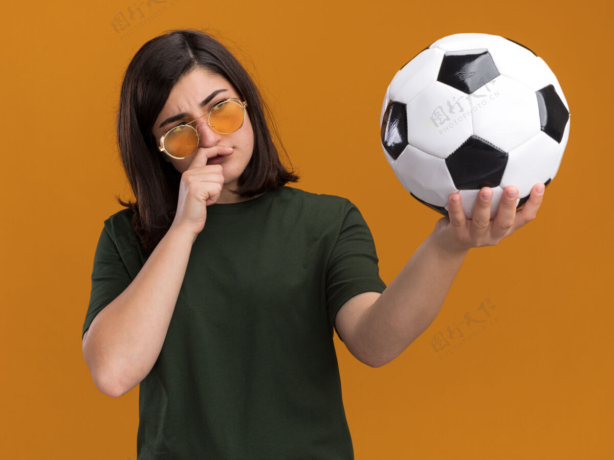 太阳体贴的年轻漂亮的高加索女孩戴着太阳眼镜把手放在下巴上拿着看球下巴女孩橙色