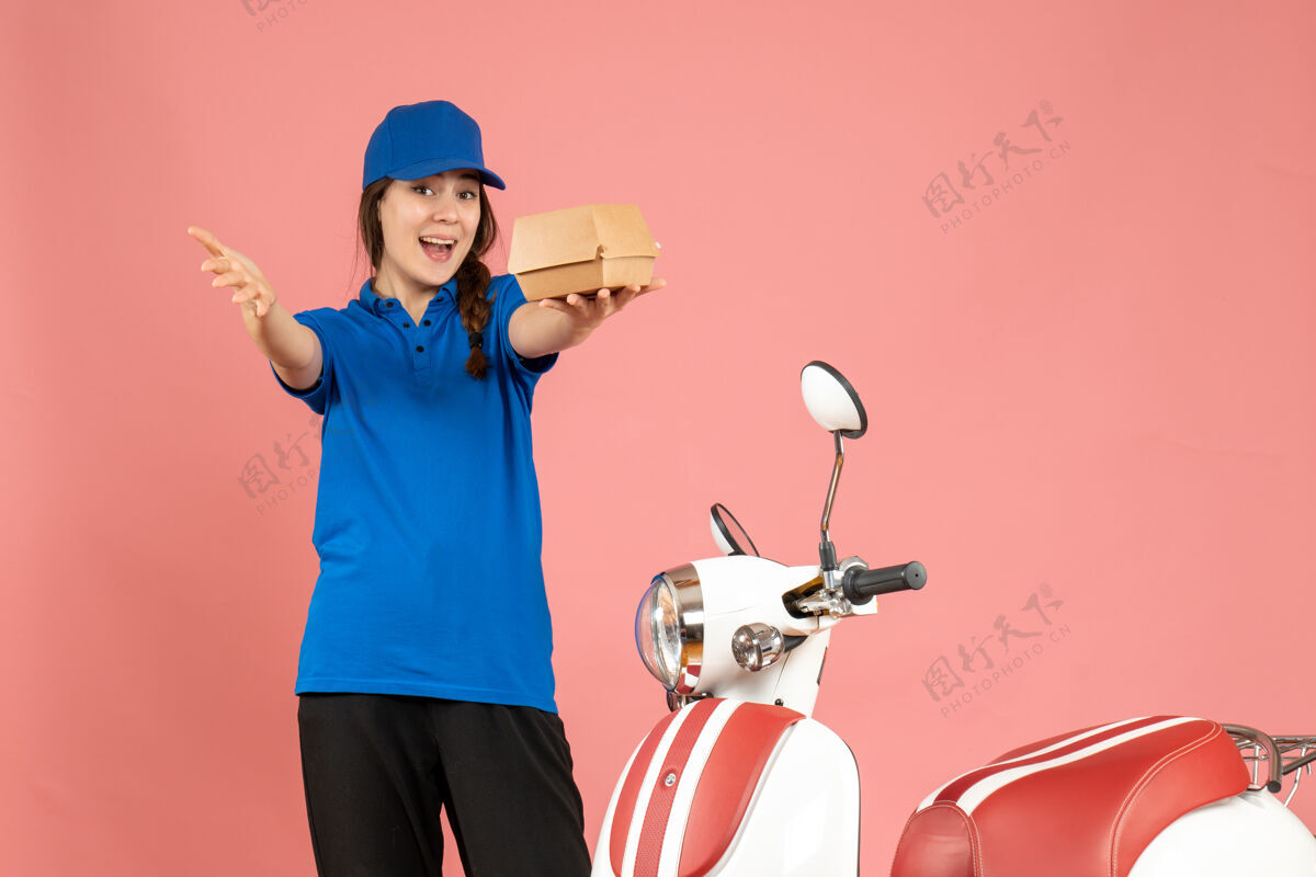 背景俯视图好奇的信使女孩站在摩托车旁边拿着蛋糕在粉彩桃色背景抱专业微笑
