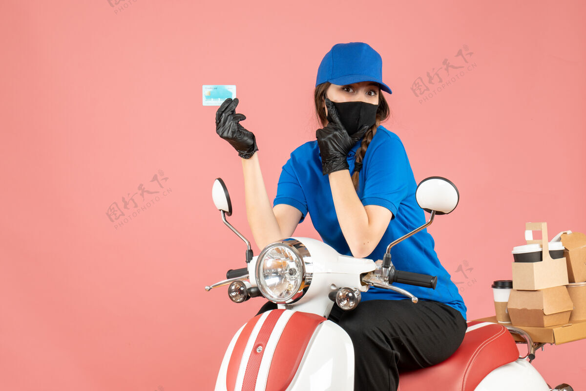 摩托车俯视图：戴着医用面罩和手套的女快递员坐在滑板车上 手里拿着银行卡 在柔和的桃色背景下递送订单车辆女快递员银行