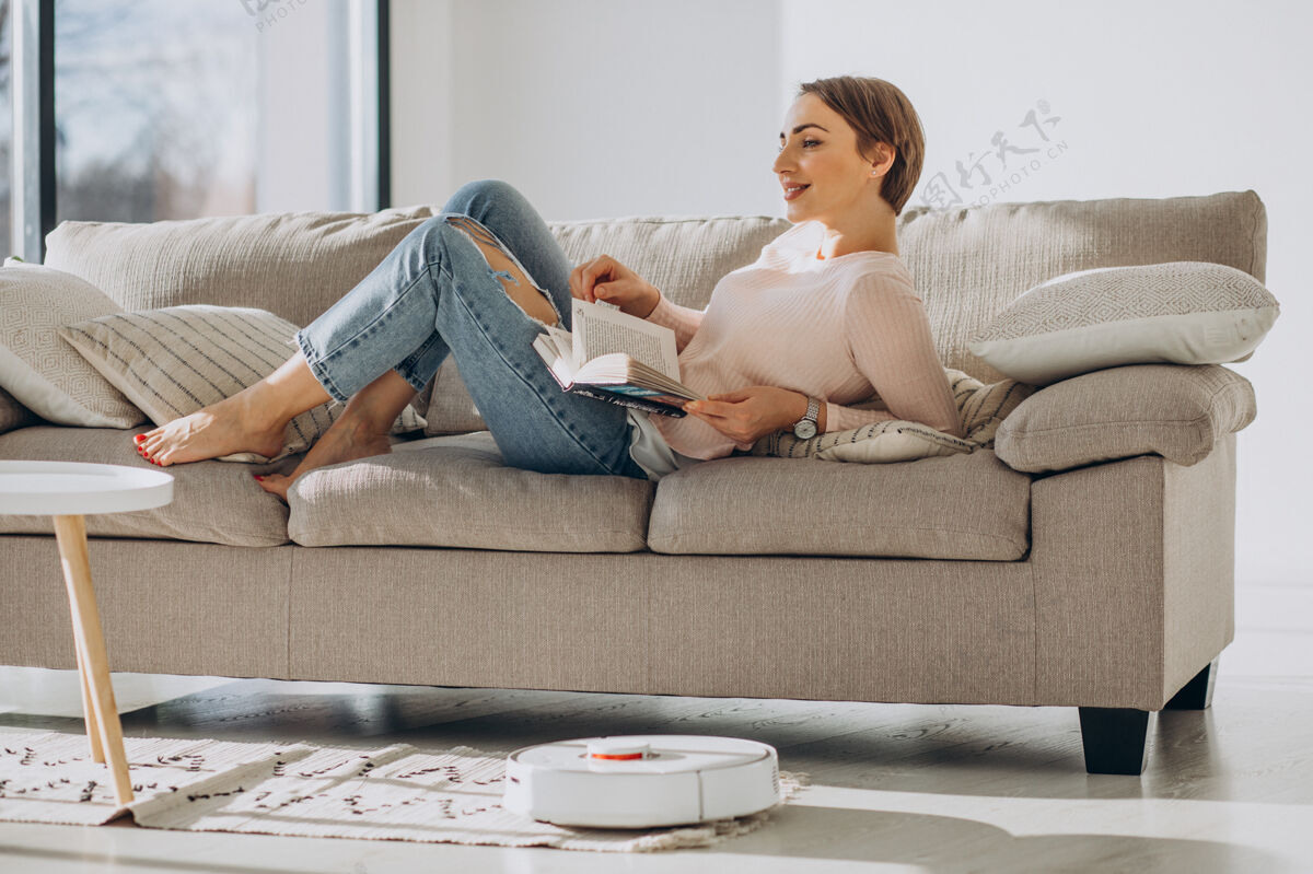 清洁年轻的女人躺在沙发上看书 而机器人吸尘器做家务吸尘器清洁家务